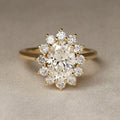 A yellow gold ring featuring a large oval center diamond surrounded by a halo of smaller round diamonds, set against a neutral textured beige background.