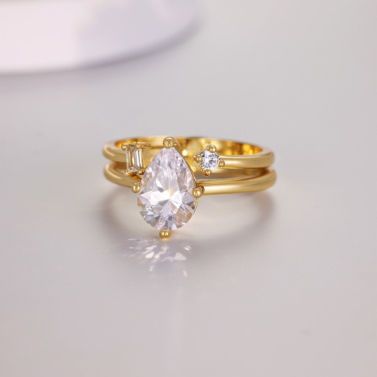 A gold ring set featuring a large pear-cut clear gemstone, a smaller baguette-cut clear gemstone, and a small round clear gemstone, displayed on a light gray reflective surface.