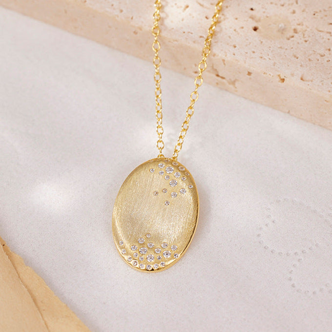 A gold oval pendant necklace with small clear rhinestones along the bottom curve, resting on a textured beige stone and white surface.