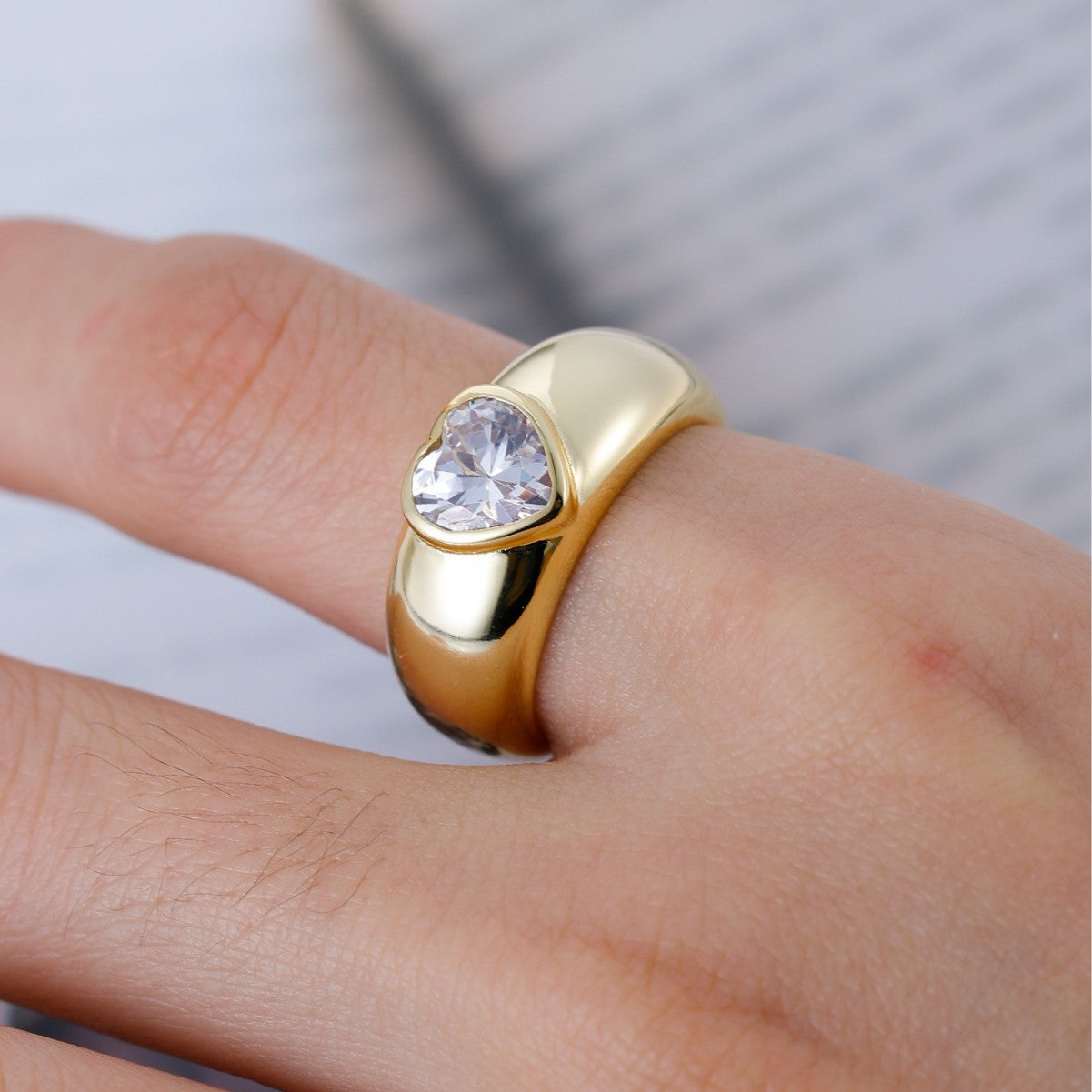 Gold ring with a clear gemstone on a finger against a neutral background