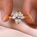A gold ring with a large oval clear center stone and two smaller marquise-cut side stones, shown being held between fingers against a soft pink fabric background.