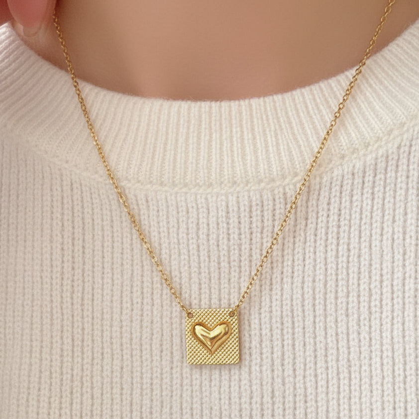 A gold necklace with a square pendant featuring a textured outer border and a smaller hollow heart in the center, worn on a person's neck over a gray ribbed top.