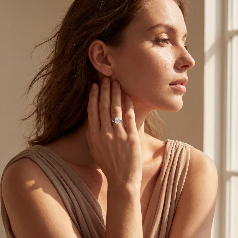 A woman in a draped taupe top wearing a silver ring with a clear center stone, shown in profile against warm window light.