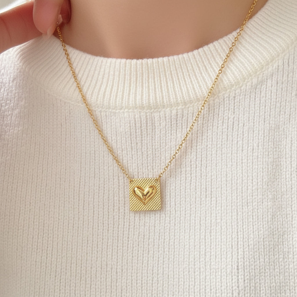 A gold necklace with a square pendant featuring a textured outer border and a smaller hollow heart in the center, worn on a person's neck over a gray ribbed top.
