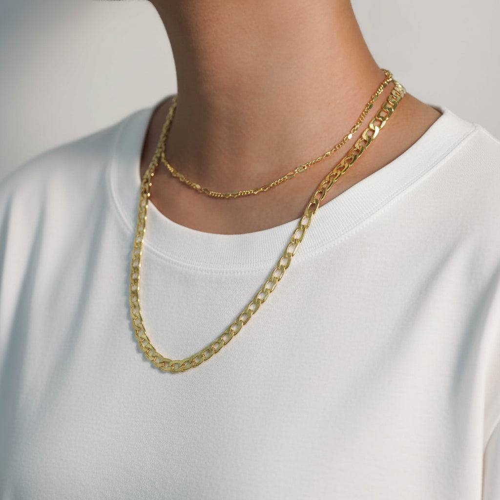 A person wearing a gray shirt with two layered gold-colored necklaces, one in solid pattern and the other in a hollow chain design.