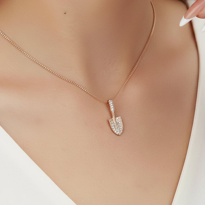 A close-up of a person wearing a delicate rose gold chain necklace with a small shovel-shaped pendant encrusted with clear stones. The necklace is shown on a model's neck against a white top.
