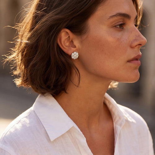 A close-up side profile of a woman with short brown hair wearing a small white flower-shaped stud earring and a white collared shirt, set against a softly blurred outdoor background.
