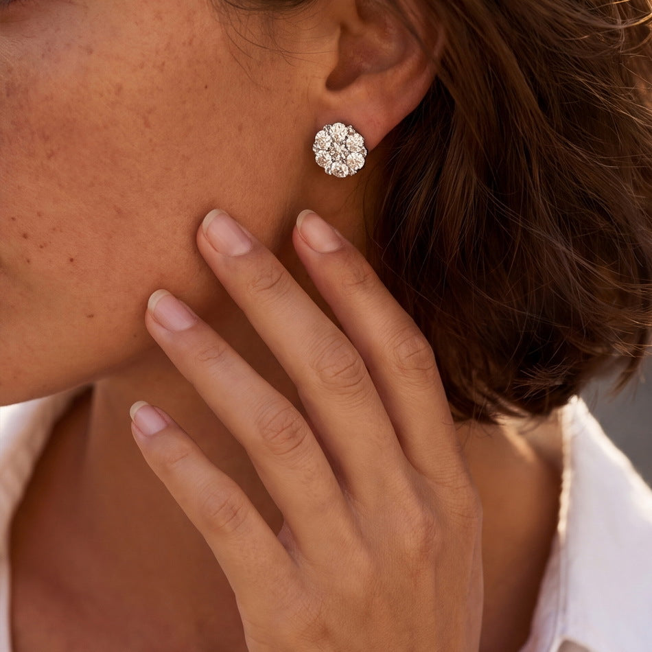 A close-up of a woman's ear wearing a small round cluster stud earring with clear stones, with her hand resting near her face.