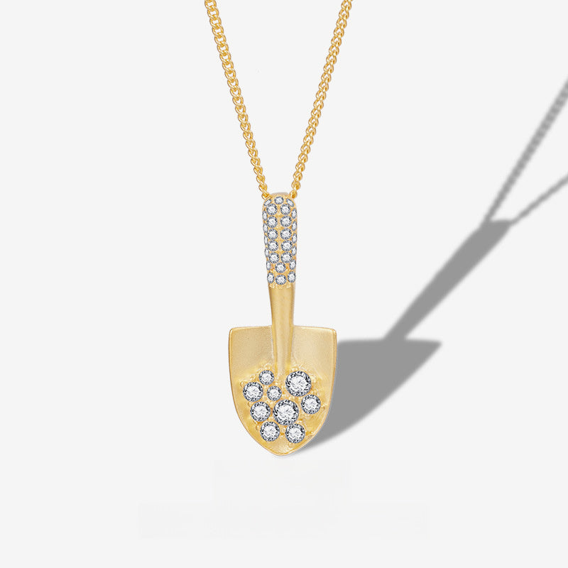 A gold shovel-shaped pendant necklace adorned with clear pavé and cluster gemstones, suspended from a gold chain against a light gray background with a soft shadow.