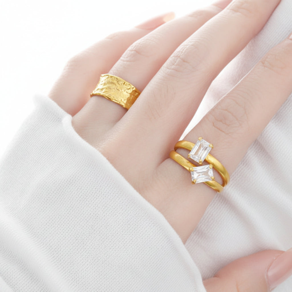 A person's hand showcasing two rings, one with a yellow gold band and the other with a silver band, each adorned with cubic zirconia stones.