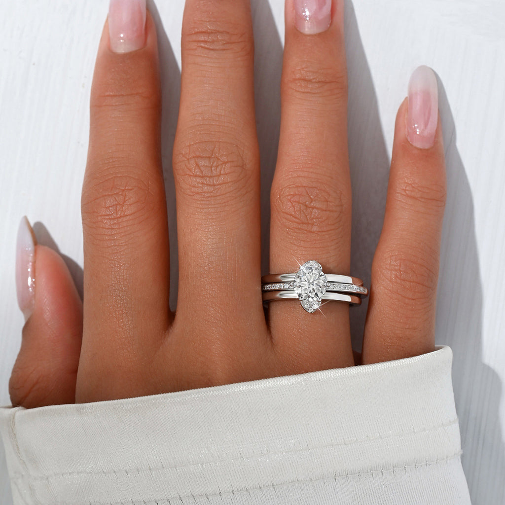 A close-up of a hand wearing a silver engagement ring with a round-cut clear center stone and a matching silver band with small pavé stones, against a white background.