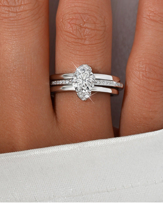 A close-up of a hand wearing a silver engagement ring with an oval-cut center stone and a matching silver band with small pavé stones, against a white background.