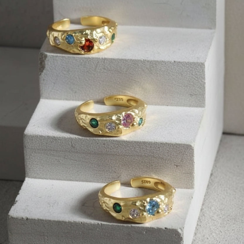 A collection of three gold-colored sterling silver adjustable rings with colorful cubic zirconia stones, displayed on a stair-like texture.