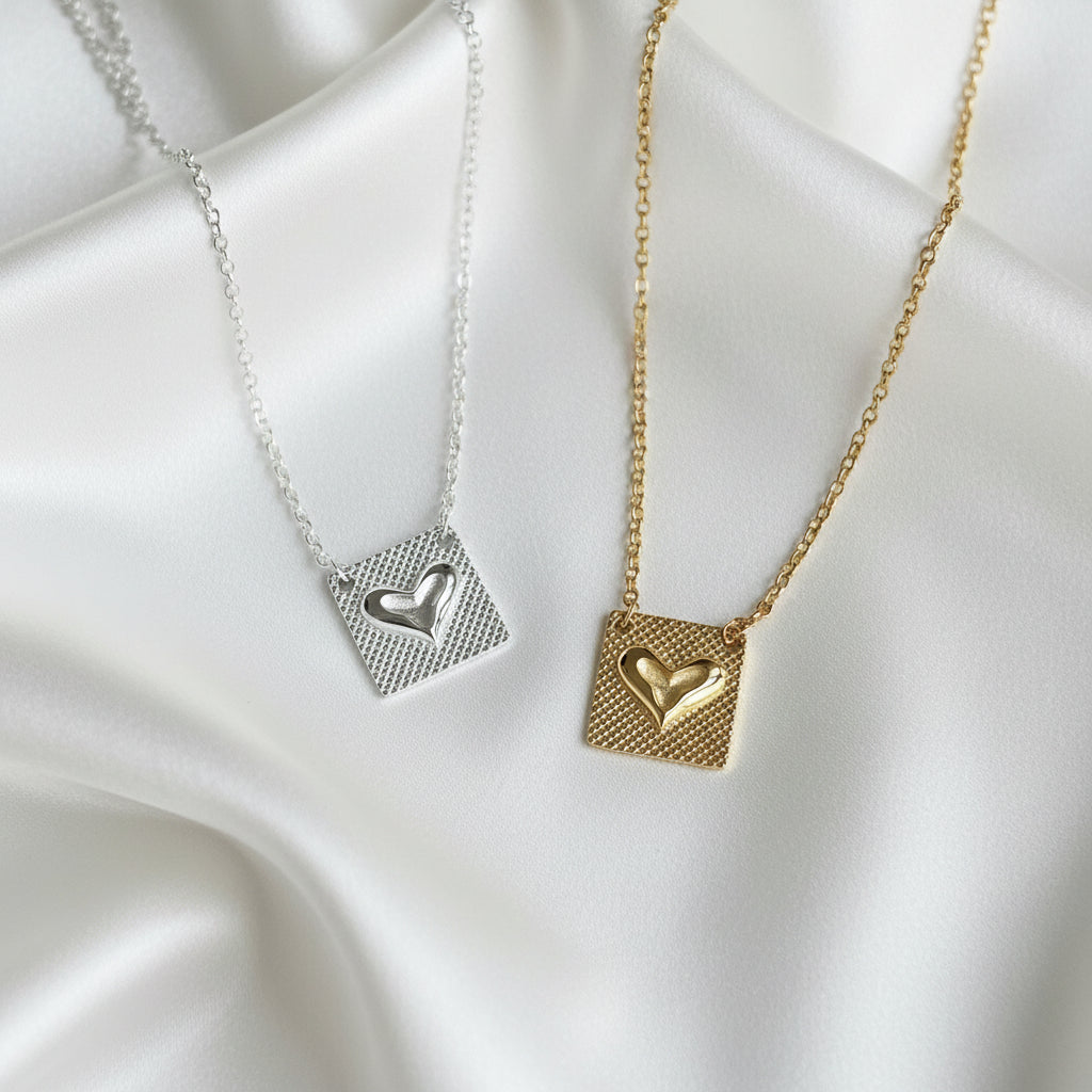 Two square pendant necklaces with textured grids and raised heart charms, shown in silver and gold finishes on a light gray textured surface.
