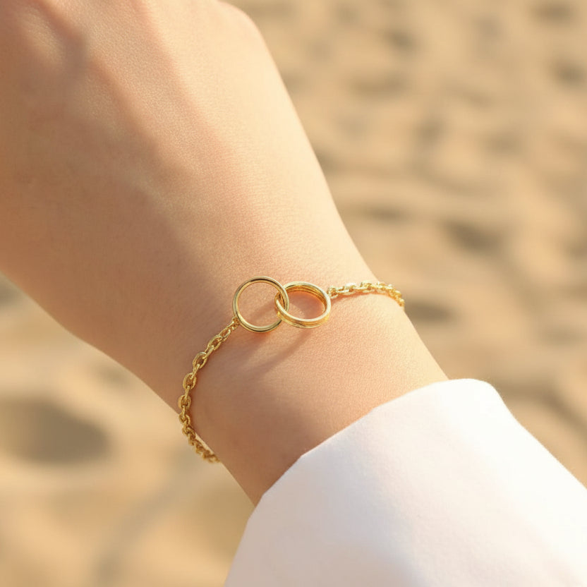 Gold bracelet with interlocking rings on a wrist, with a blurred background of a book and wooden surface.