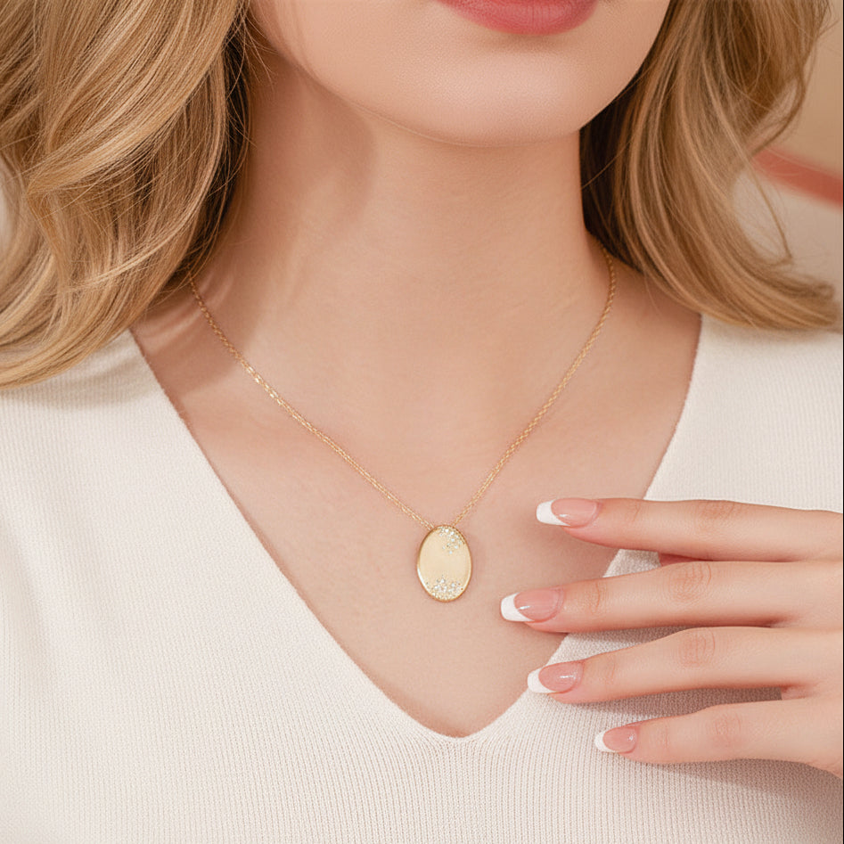 A close-up of a woman wearing a delicate gold chain necklace with an oval pendant featuring a textured, pearl-like center, styled with a cream-colored top.